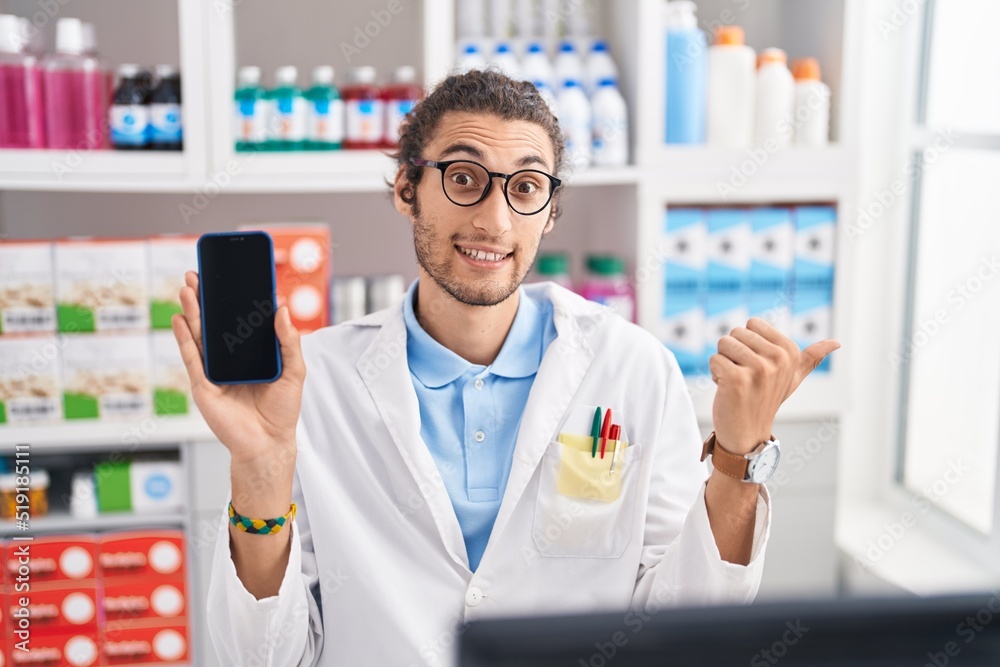 Young hispanic man working at pharmacy drugstore showing smartphone screen pointing thumb up to the side smiling happy with open mouth
