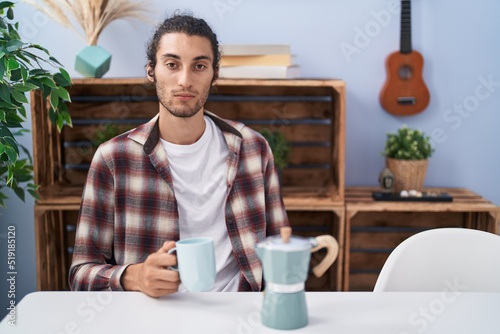Young hispanic man drinking coffee from french coffee maker thinking attitude and sober expression looking self confident