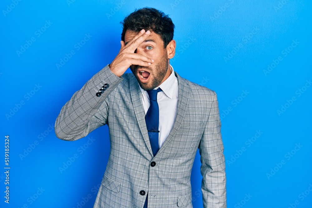 Handsome man with beard wearing business suit and tie peeking in shock covering face and eyes with hand, looking through fingers with embarrassed expression.