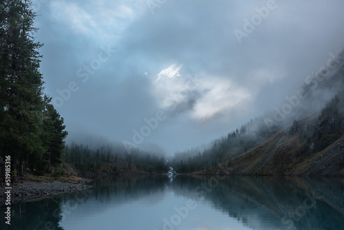 Tranquil scenery with snow castle in clouds. Mountain creek flows from forest hills into glacial lake. Snowy mountains in fog clearance. Small river and coniferous trees reflected in calm alpine lake.