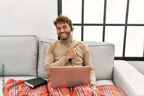 Young handsome man with beard wearing operator headset working from home cheerful with a smile on face pointing with hand and finger up to the side with happy and natural expression