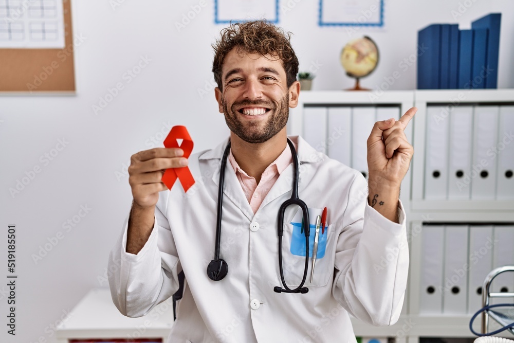 Young hispanic doctor man holding support red ribbon at medical clinic smiling happy pointing with hand and finger to the side Young hispanic doctor man holding support red ribbon at medical clinic smiling happy pointing with hand and finger to the side