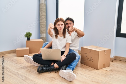 Young couple using laptop at new home looking unhappy and angry showing rejection and negative with thumbs down gesture. bad expression.