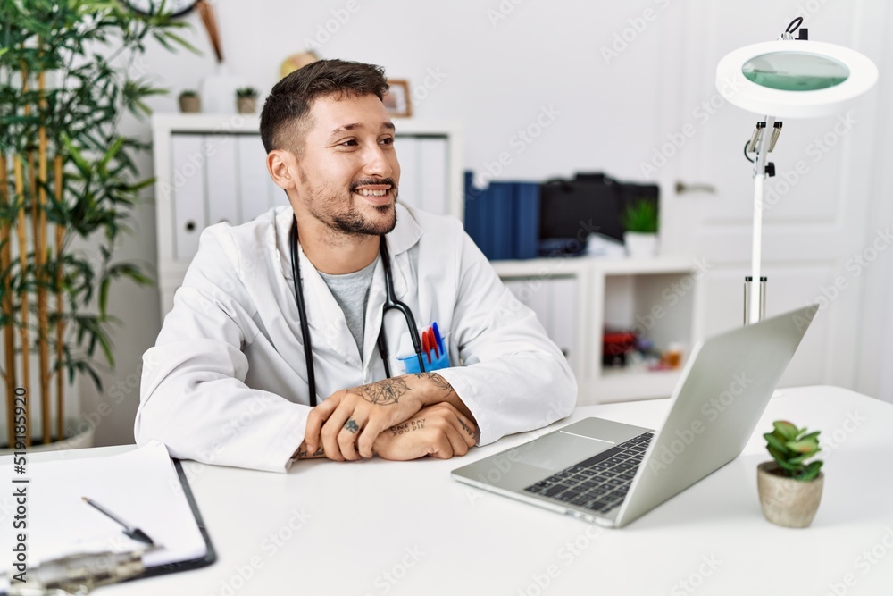 Young doctor working at the clinic using computer laptop looking away to side with smile on face, natural expression. laughing confident.