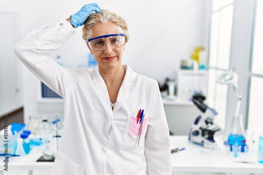 Middle age blonde woman working at scientist laboratory confuse and wonder about question. uncertain with doubt, thinking with hand on head. pensive concept.