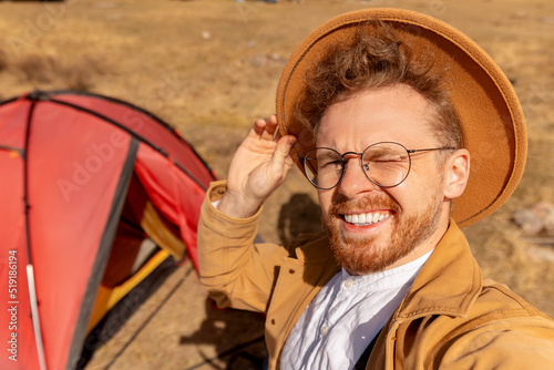 Happy hikker man with hat making selfie photo portrait background red tent and mountains photo