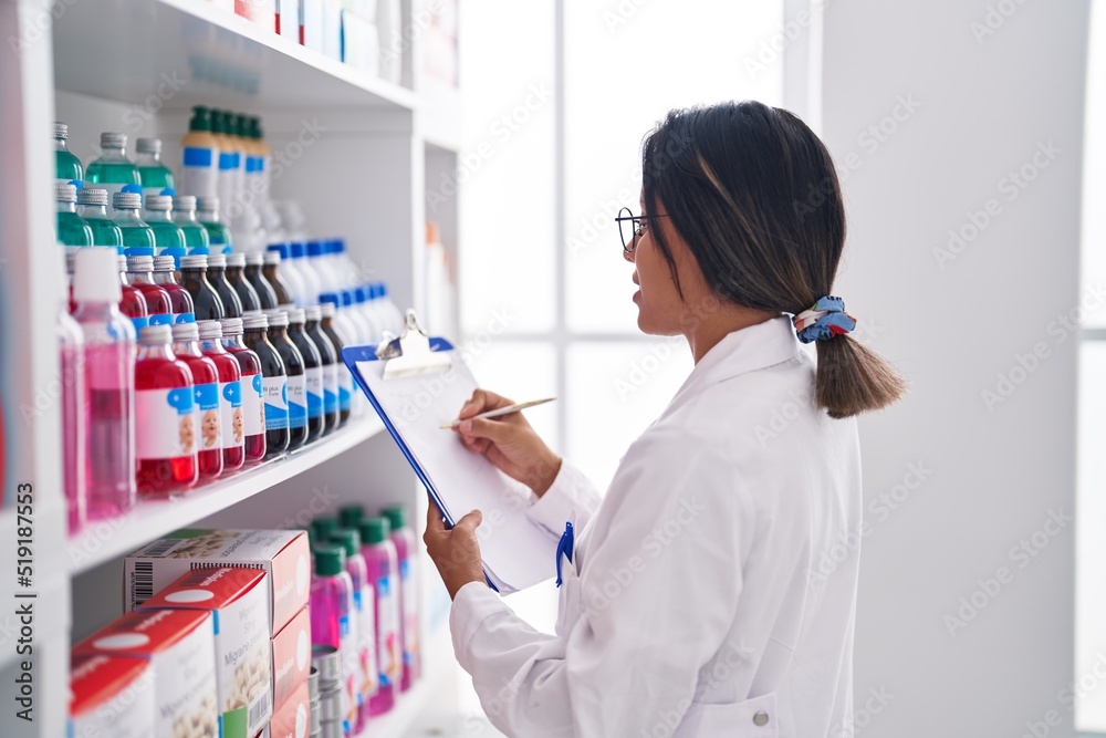 Young hispanic woman pharmacist writing on document looking shelving at pharmacy Young hispanic woman pharmacist writing on document looking shelving at pharmacy