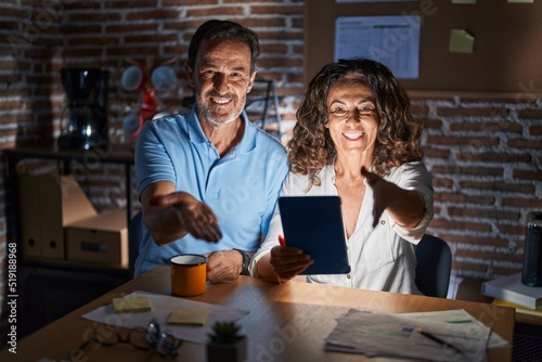 Middle age hispanic couple using touchpad sitting on the table at night smiling cheerful offering palm hand giving assistance and acceptance.