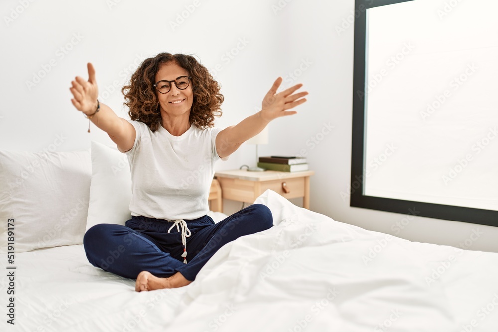 Middle age hispanic woman sitting on the bed at home looking at the camera smiling with open arms for hug. cheerful expression embracing happiness. Middle age hispanic woman sitting on the bed at home looking at the camera smiling with open arms for hug. cheerful expression embracing happiness.