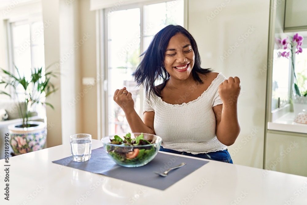 Young hispanic woman eating healthy salad at home very happy and excited doing winner gesture with arms raised, smiling and screaming for success. celebration concept. Young hispanic woman eating healthy salad at home very happy and excited doing winner gesture with arms raised, smiling and screaming for success. celebration concept.