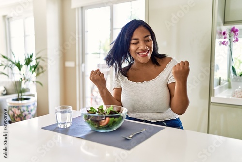 Young hispanic woman eating healthy salad at home very happy and excited doing winner gesture with arms raised  smiling and screaming for success. celebration concept.