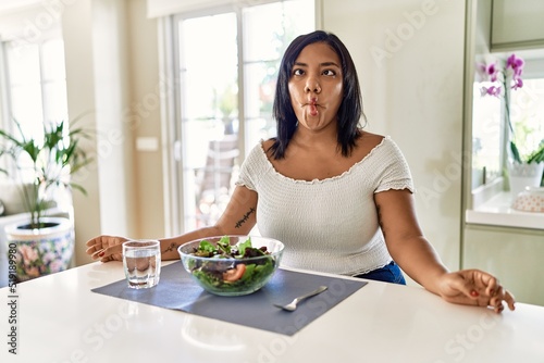 Young hispanic woman eating healthy salad at home making fish face with lips  crazy and comical gesture. funny expression.