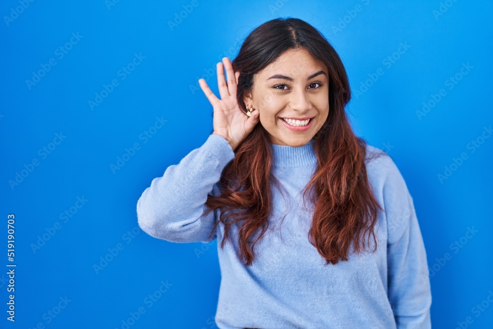 Hispanic young woman standing over blue background smiling with hand over ear listening an hearing to rumor or gossip. deafness concept. Hispanic young woman standing over blue background smiling with hand over ear listening an hearing to rumor or gossip. deafness concept.