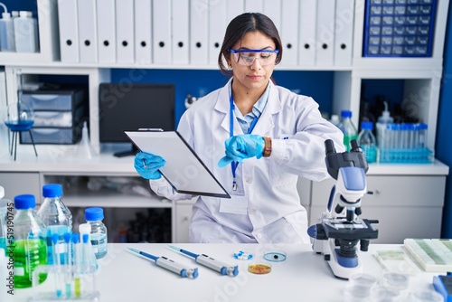 Hispanic young woman working at scientist laboratory checking the time on wrist watch  relaxed and confident
