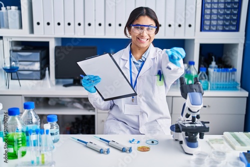 Hispanic young woman working at scientist laboratory pointing to you and the camera with fingers  smiling positive and cheerful