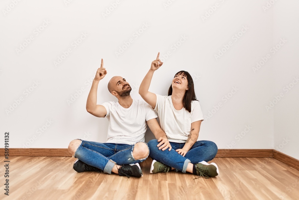 Young hispanic couple smiling happy pointing with finger to the ceiling at empty new home.