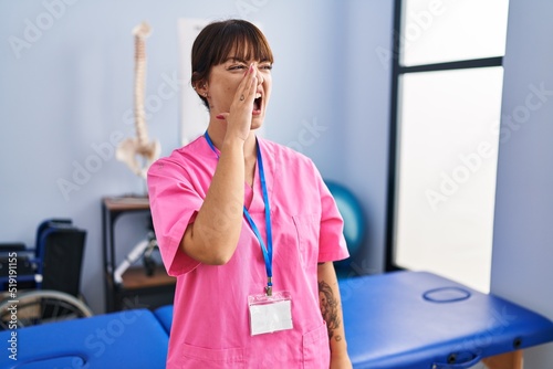 Young brunette woman working at rehabilitation clinic shouting and screaming loud to side with hand on mouth. communication concept.