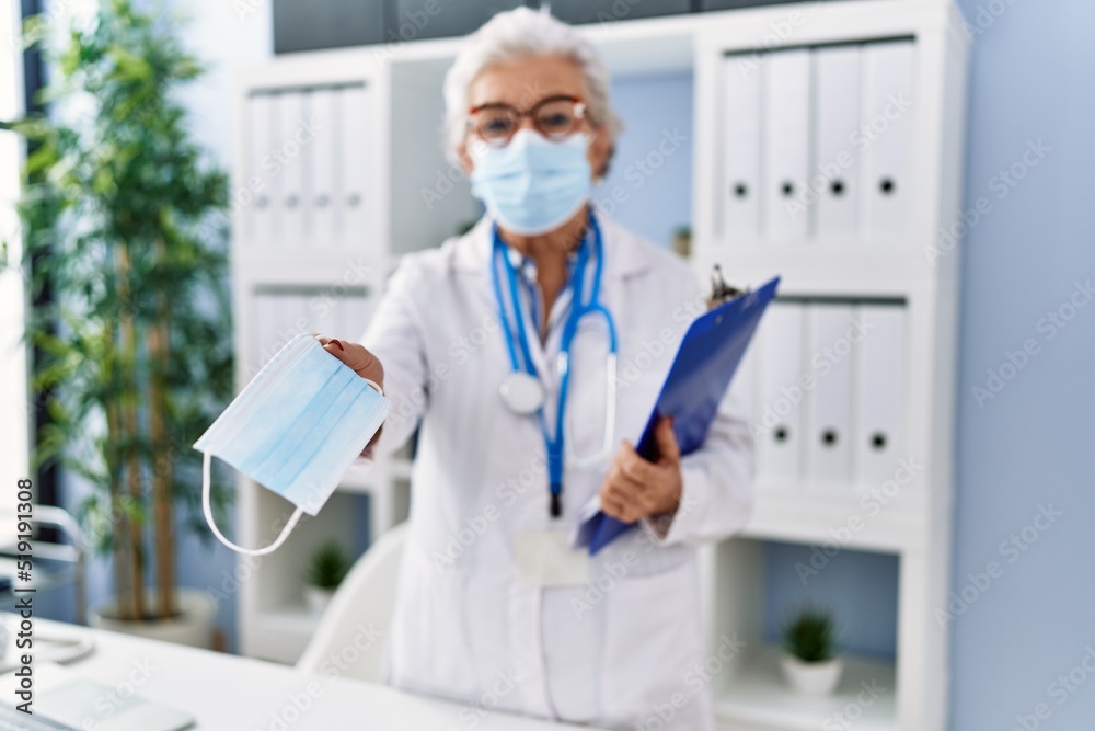 Senior grey-haired woman wearing doctor uniform giving medical mask at clinic Senior grey-haired woman wearing doctor uniform giving medical mask at clinic