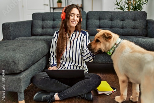 Young hispanic woman using laptop sitting on floor with dog at home