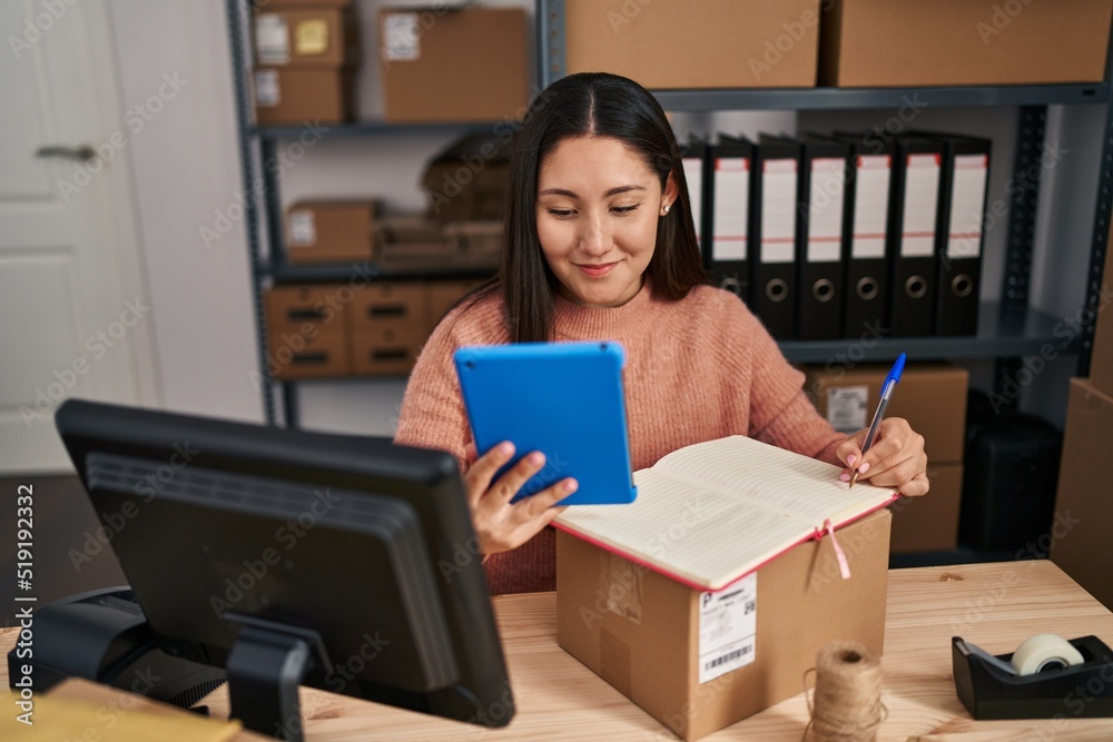 Young hispanic woman ecommerce business worker writing on notebook and using touchpad at office Young hispanic woman ecommerce business worker writing on notebook and using touchpad at office