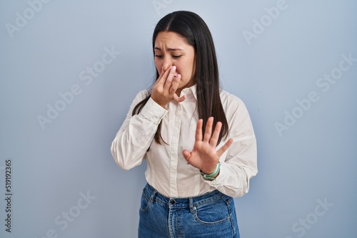 Foto Young latin woman standing over blue background smelling something stinky and disgusting, intolerable smell, holding breath with fingers on nose