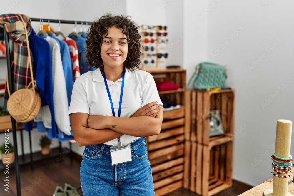 Fototapeta premium Young latin shopkeeper woman smiling happy standing with arms crossed gesture at clothing store.