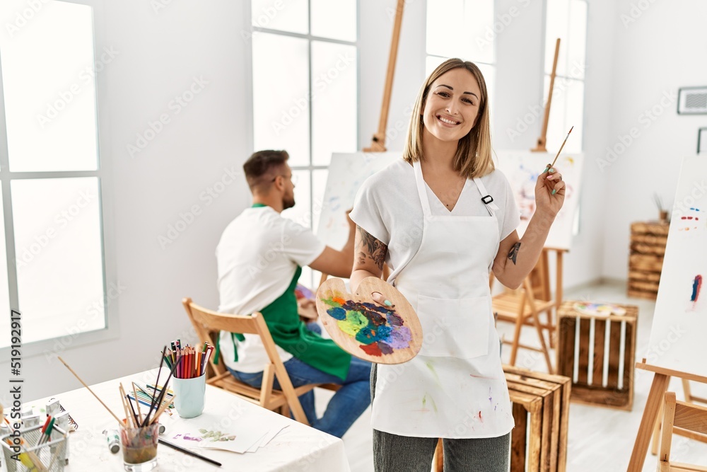 Young caucasian couple drawing at art studio. Girl smiling happy standing and holding paintbrush and palette. Young caucasian couple drawing at art studio. Girl smiling happy standing and holding paintbrush and palette.