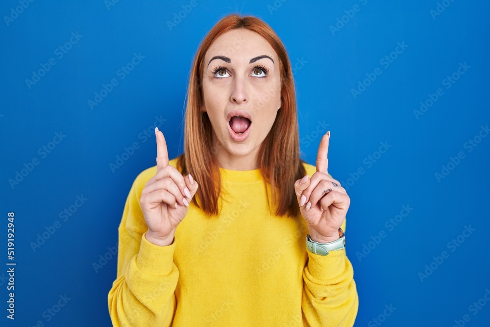 Young woman standing over blue background amazed and surprised looking up and pointing with fingers and raised arms. Young woman standing over blue background amazed and surprised looking up and pointing with fingers and raised arms.