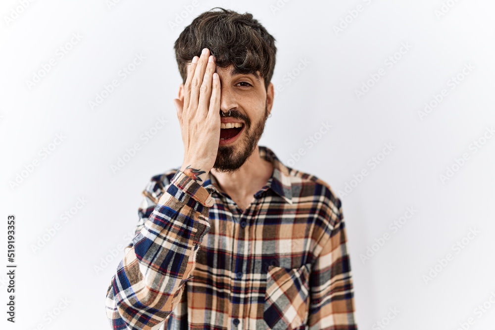 Hispanic man with beard standing over isolated background covering one eye with hand, confident smile on face and surprise emotion. Hispanic man with beard standing over isolated background covering one eye with hand, confident smile on face and surprise emotion.