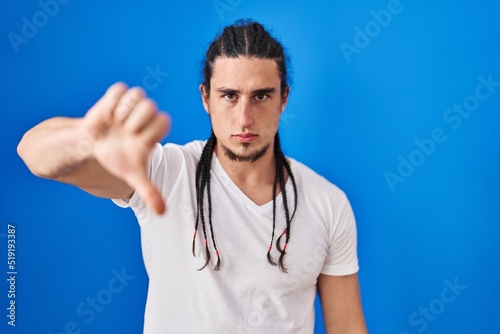 Hispanic man with long hair standing over blue background looking unhappy and angry showing rejection and negative with thumbs down gesture. bad expression.