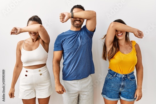 Group of young hispanic people standing over isolated background covering eyes with arm smiling cheerful and funny. blind concept.