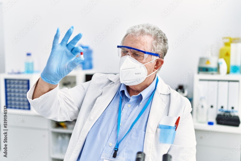 Middle age grey-haired man wearing scientist uniform hoding pill at laboratory Middle age grey-haired man wearing scientist uniform hoding pill at laboratory