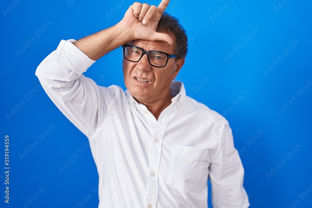 Naklejka premium Middle age hispanic man standing over blue background making fun of people with fingers on forehead doing loser gesture mocking and insulting.