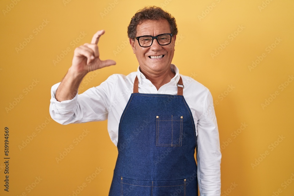 Middle age hispanic man wearing professional cook apron smiling and confident gesturing with hand doing small size sign with fingers looking and the camera. measure concept. Middle age hispanic man wearing professional cook apron smiling and confident gesturing with hand doing small size sign with fingers looking and the camera. measure concept.