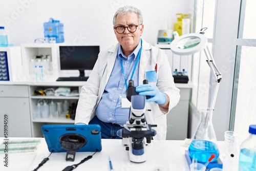 Middle age grey-haired man wearing scientist uniform using microscope and touchpad at laboratory