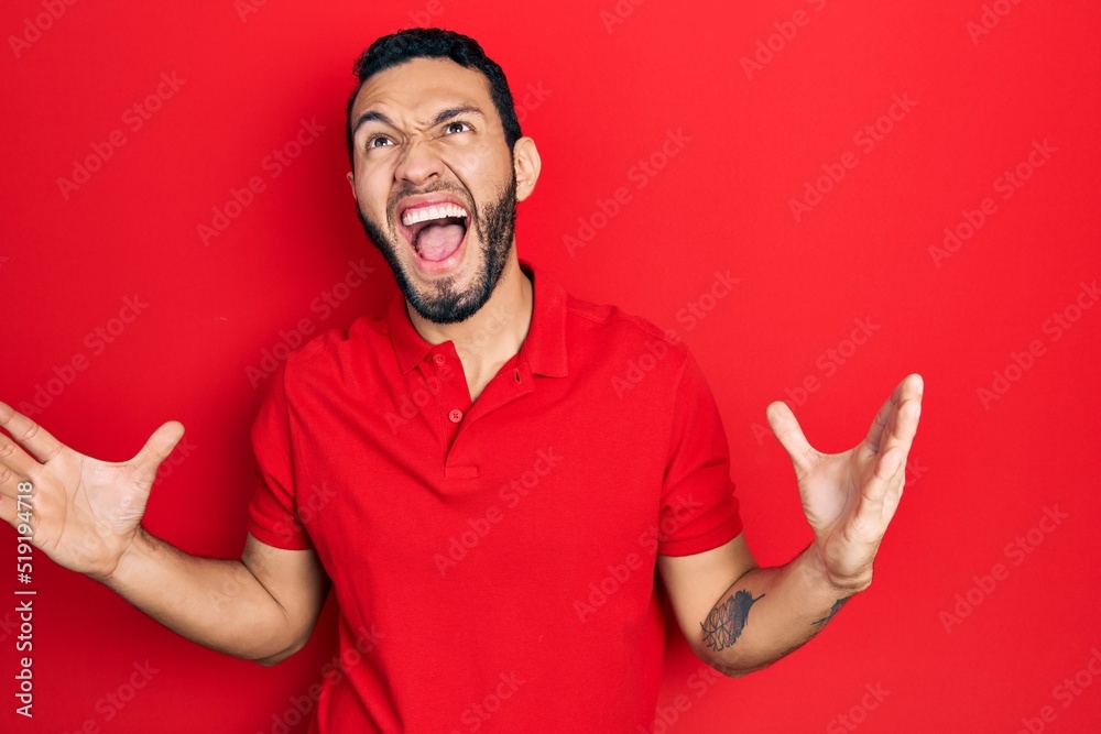 Hispanic man with beard wearing casual red t shirt crazy and mad shouting and yelling with aggressive expression and arms raised. frustration concept. Hispanic man with beard wearing casual red t shirt crazy and mad shouting and yelling with aggressive expression and arms raised. frustration concept.
