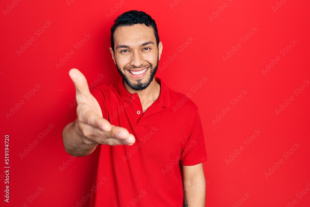 Hispanic man with beard wearing casual red t shirt smiling friendly offering handshake as greeting and welcoming. successful business. Hispanic man with beard wearing casual red t shirt smiling friendly offering handshake as greeting and welcoming. successful business.
