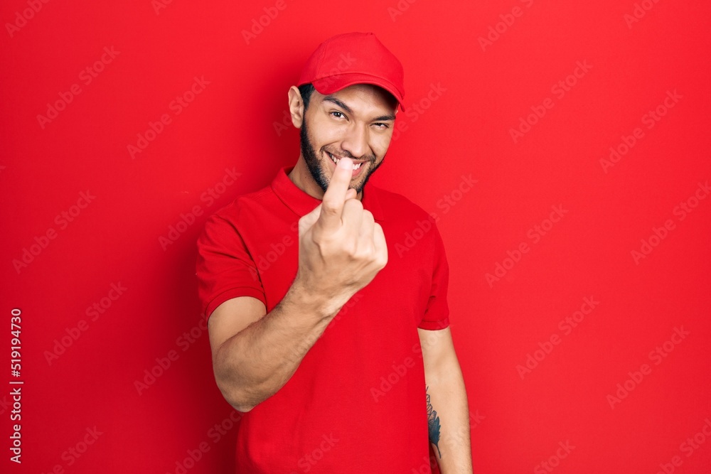 Hispanic man with beard wearing delivery uniform and cap beckoning come here gesture with hand inviting welcoming happy and smiling