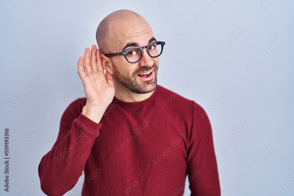 Young bald man with beard standing over white background wearing glasses smiling with hand over ear listening an hearing to rumor or gossip. deafness concept. Young bald man with beard standing over white background wearing glasses smiling with hand over ear listening an hearing to rumor or gossip. deafness concept.