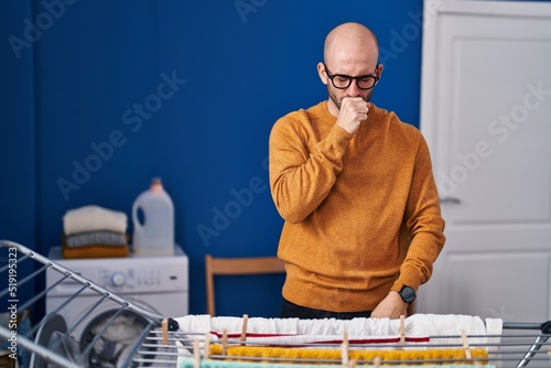 Young bald man with beard hanging clothes at clothesline feeling unwell and coughing as symptom for cold or bronchitis. health care concept.