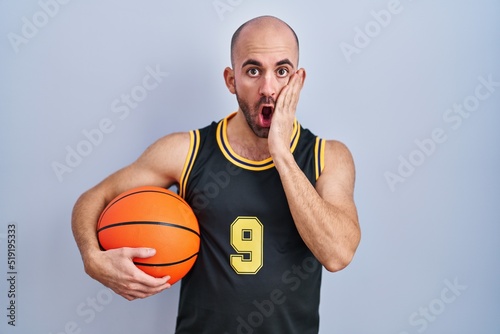 Young bald man with beard wearing basketball uniform holding ball afraid and shocked, surprise and amazed expression with hands on face