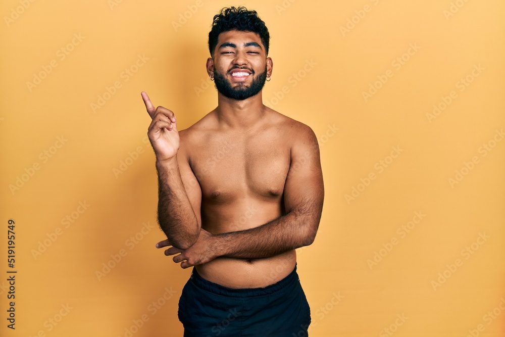 Φωτογραφία Arab man with beard wearing swimwear shirtless with a big smile on face, pointing with hand and finger to the side looking at the camera