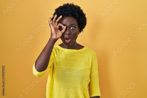 African young woman standing over yellow studio doing ok gesture shocked with surprised face, eye looking through fingers. unbelieving expression. © Krakenimages.com