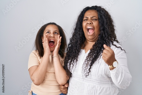 Mother and young daughter standing over white background shouting angry out loud with hands over mouth