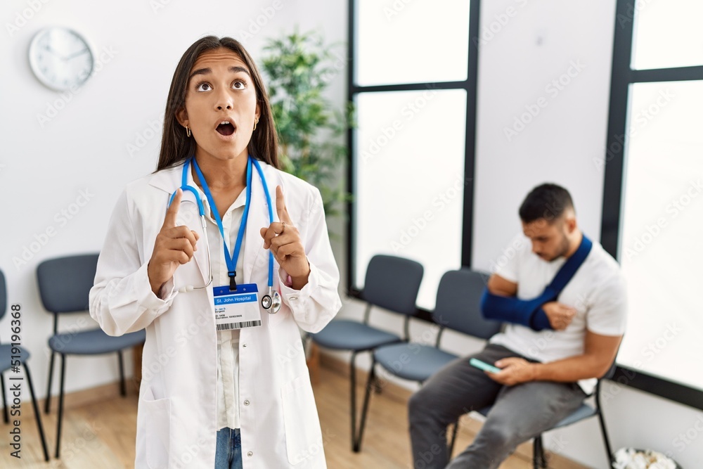 Young asian doctor woman at waiting room with a man with a broken arm amazed and surprised looking up and pointing with fingers and raised arms. Young asian doctor woman at waiting room with a man with a broken arm amazed and surprised looking up and pointing with fingers and raised arms.