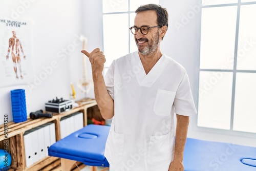 Middle age man with beard working at pain recovery clinic smiling with happy face looking and pointing to the side with thumb up.