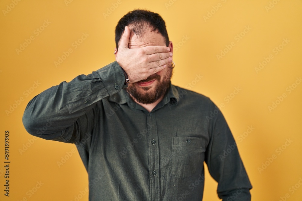 Plus size hispanic man with beard standing over yellow background smiling and laughing with hand on face covering eyes for surprise. blind concept. Plus size hispanic man with beard standing over yellow background smiling and laughing with hand on face covering eyes for surprise. blind concept.