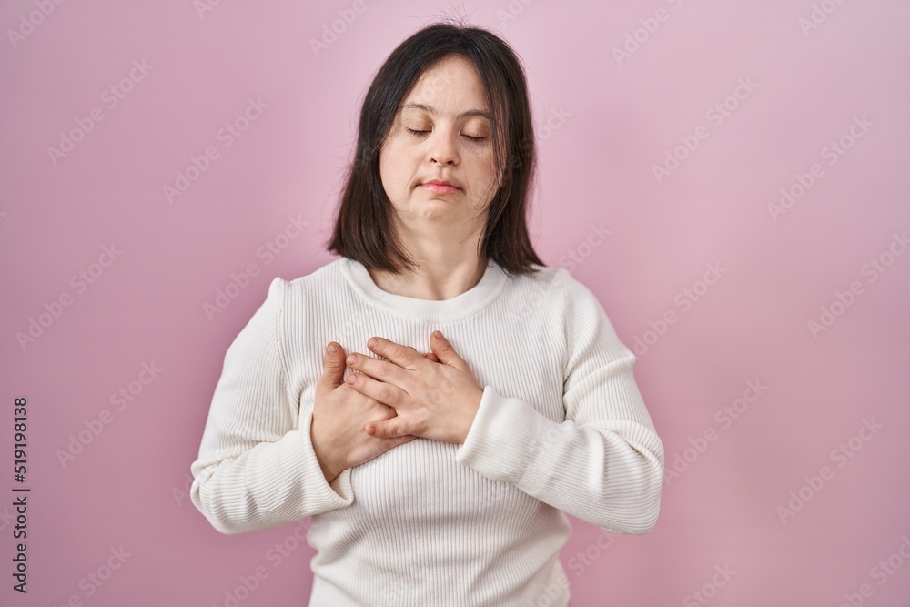 Woman with down syndrome standing over pink background smiling with hands on chest with closed eyes and grateful gesture on face. health concept. Woman with down syndrome standing over pink background smiling with hands on chest with closed eyes and grateful gesture on face. health concept.