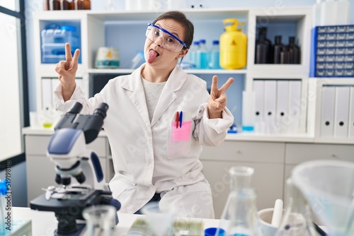 Hispanic girl with down syndrome working at scientist laboratory smiling with tongue out showing fingers of both hands doing victory sign. number two.