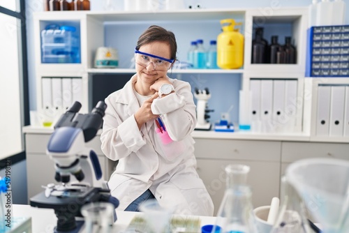 Hispanic girl with down syndrome working at scientist laboratory in hurry pointing to watch time  impatience  looking at the camera with relaxed expression